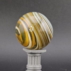 May include: A large, swirled glass marble with shades of yellow, brown, and white, resting on a small, gray, fluted column. The marble has a glossy finish and is the focal point of the image.