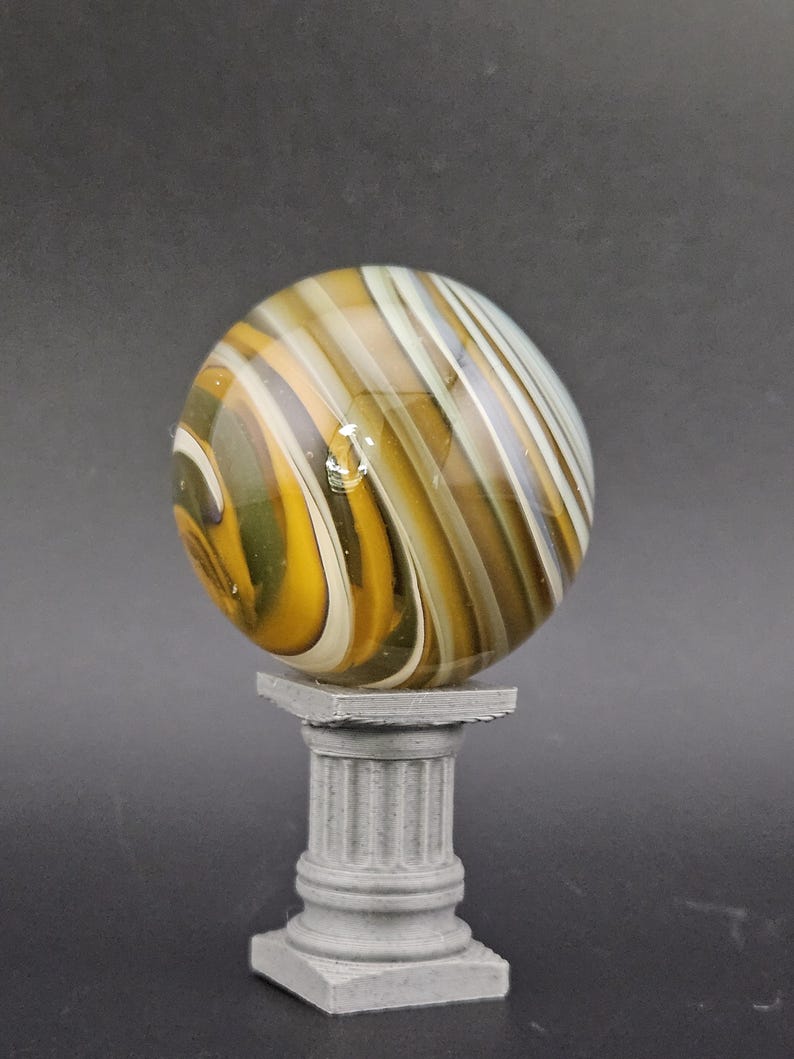 May include: A large, swirled glass marble with shades of yellow, brown, and white, resting on a small, gray, fluted column. The marble has a glossy finish and is the focal point of the image.