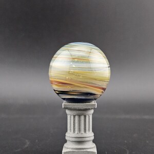 May include: A decorative glass sphere with horizontal stripes in shades of yellow, orange, and blue, resting on a small, gray, classical-style pedestal. The sphere has a glossy finish and appears to be made of layered glass.