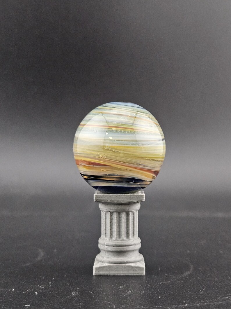 May include: A decorative glass sphere with horizontal stripes in shades of yellow, orange, and blue, resting on a small, gray, classical-style pedestal. The sphere has a glossy finish and appears to be made of layered glass.