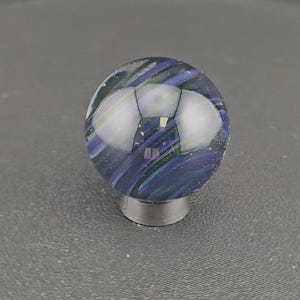 May include: A spherical glass marble featuring swirling patterns of deep blue, purple, and dark green hues. The surface is smooth and reflective, showing light highlights. The marble is supported by a small, circular, dark gray metallic stand.