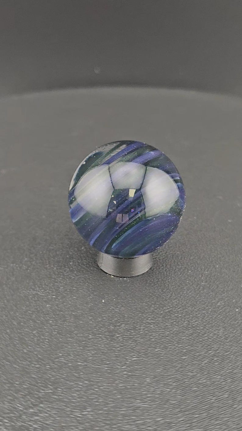 May include: A spherical glass marble featuring swirling patterns of deep blue, purple, and dark green hues. The surface is smooth and reflective, showing light highlights. The marble is supported by a small, circular, dark gray metallic stand.