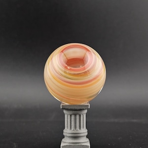 May include: A decorative glass orb with swirling bands of orange, yellow, and red, resting on a small, gray, classical-style pedestal. The orb has a hollowed-out center. The background is a solid black.