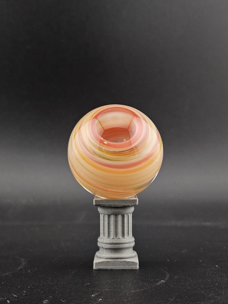 May include: A decorative glass orb with swirling bands of orange, yellow, and red, resting on a small, gray, classical-style pedestal. The orb has a hollowed-out center. The background is a solid black.