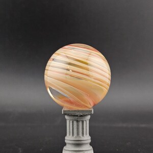 May include: A decorative glass sphere with swirling orange, yellow, and white colors, resting on a small, gray, classical-style pedestal. The sphere has a polished, smooth surface. The pedestal has a square base and a fluted column.