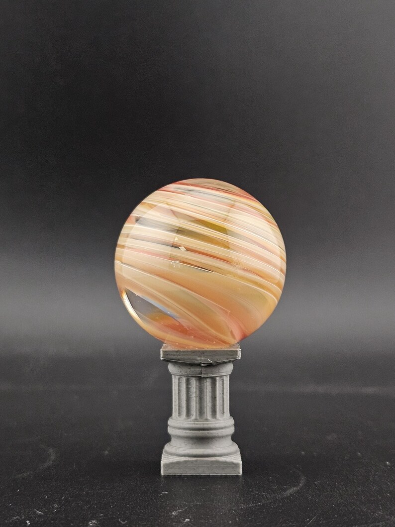 May include: A decorative glass sphere with swirling orange, yellow, and white colors, resting on a small, gray, classical-style pedestal. The sphere has a polished, smooth surface. The pedestal has a square base and a fluted column.