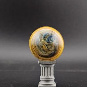 May include: A decorative glass orb with yellow, blue, and brown swirls, mounted on a small, gray, classical-style column. The orb has a glossy finish and sits centered on the column, creating a unique display piece.