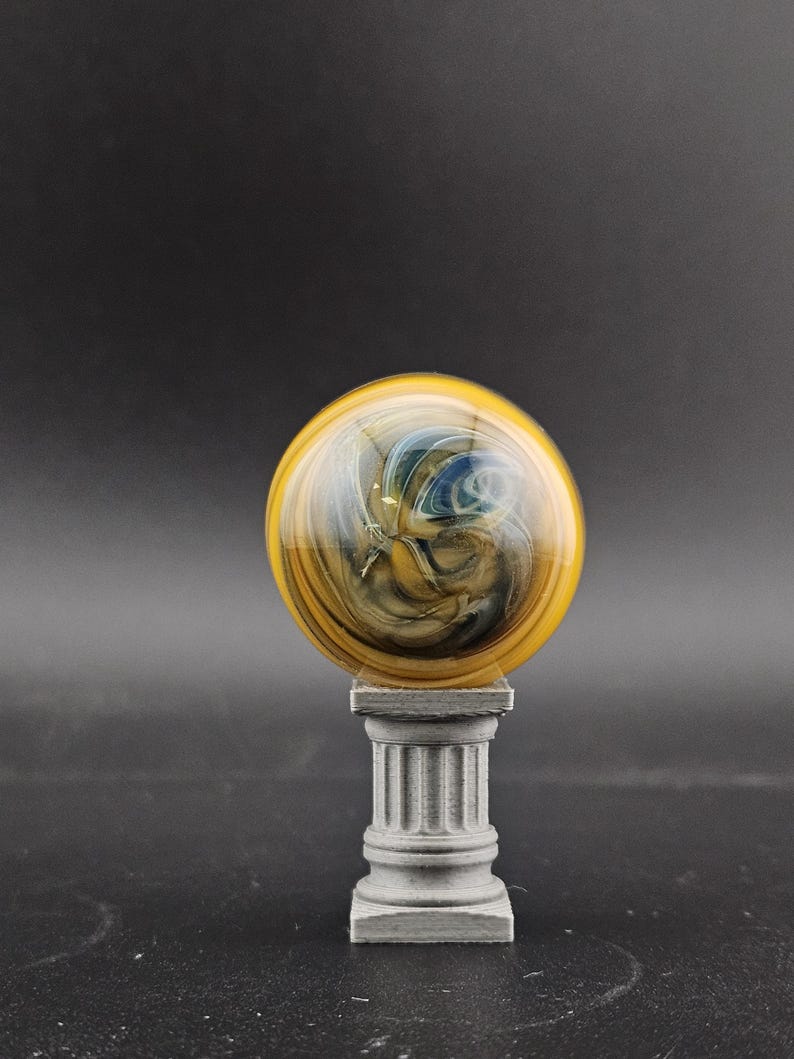May include: A decorative glass orb with yellow, blue, and brown swirls, mounted on a small, gray, classical-style column. The orb has a glossy finish and sits centered on the column, creating a unique display piece.
