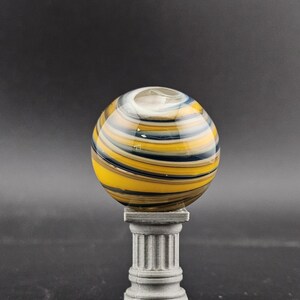 May include: A decorative glass orb with yellow, black, and blue stripes, resting on a small, gray, classical-style pedestal. The orb has a clear opening at the top. The background is a solid dark gray.