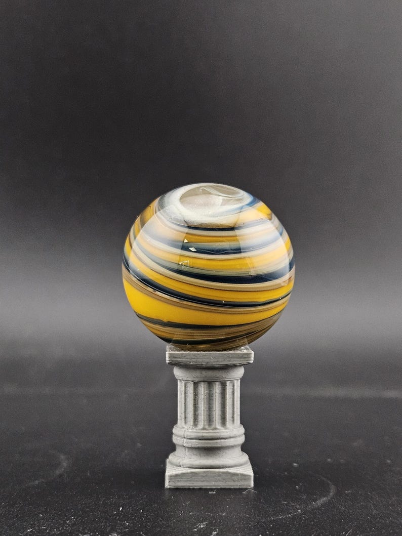 May include: A decorative glass orb with yellow, black, and blue stripes, resting on a small, gray, classical-style pedestal. The orb has a clear opening at the top. The background is a solid dark gray.