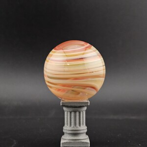 May include: A decorative glass orb with swirling orange, yellow, and white colors, resting on a gray, fluted pedestal. The orb is spherical and appears to be made of layered glass. The pedestal has a classic column design.
