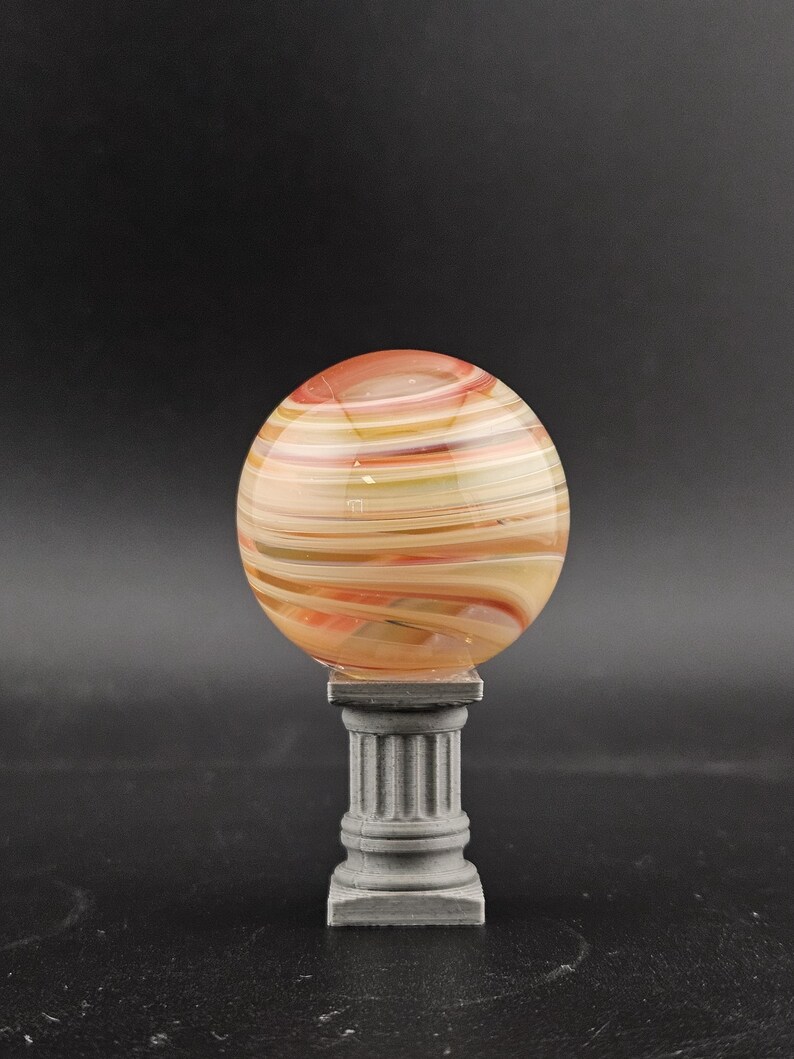 May include: A decorative glass orb with swirling orange, yellow, and white colors, resting on a gray, fluted pedestal. The orb is spherical and appears to be made of layered glass. The pedestal has a classic column design.