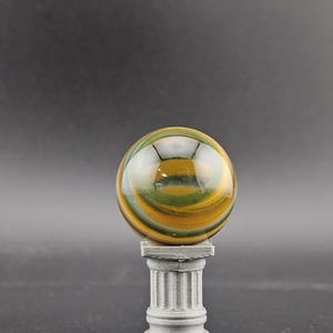 May include: A polished, spherical gemstone with swirling bands of green, yellow, and brown, resting atop a small, gray, fluted column. The orb reflects light, creating a glossy appearance. The column has a square base and a detailed, classical design.