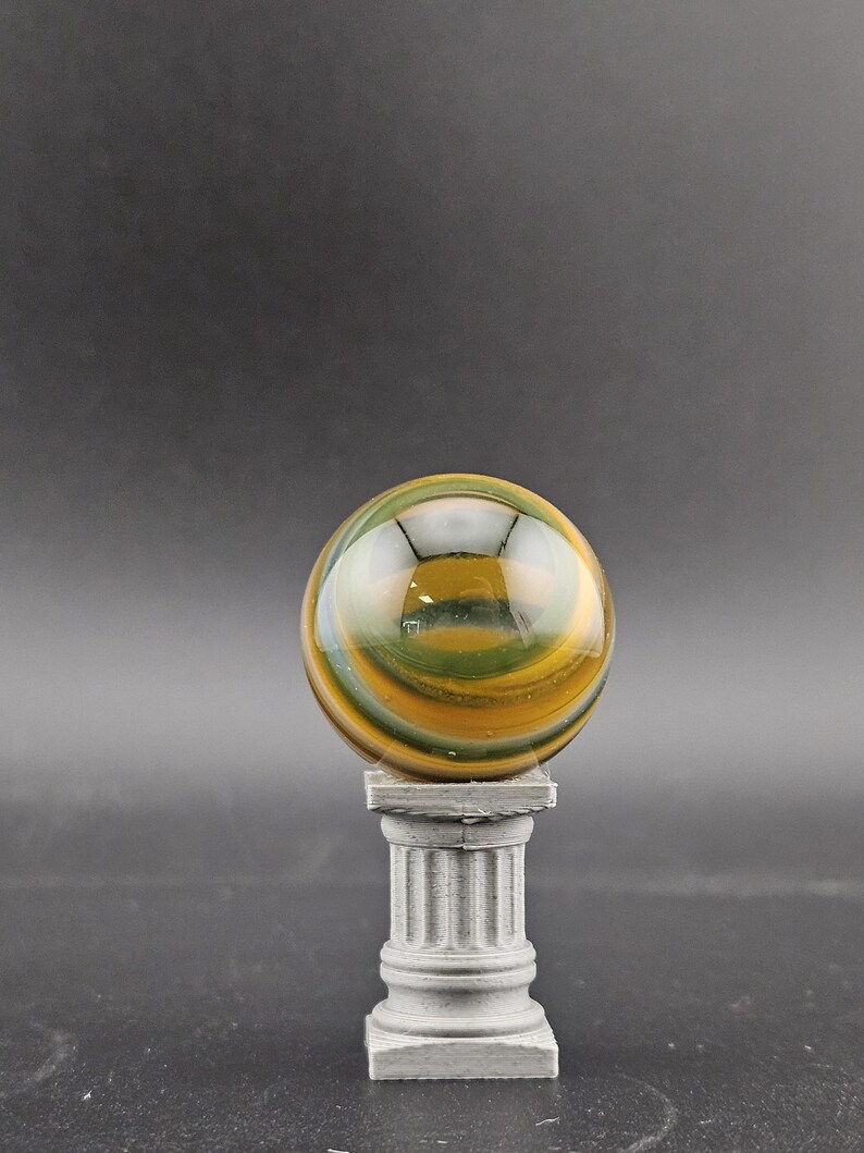 May include: A polished, spherical gemstone with swirling bands of green, yellow, and brown, resting atop a small, gray, fluted column. The orb reflects light, creating a glossy appearance. The column has a square base and a detailed, classical design.