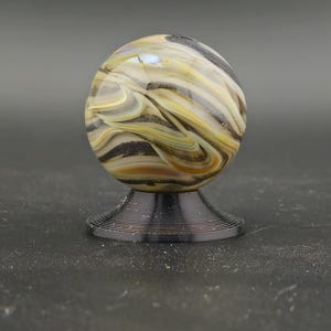 May include: A decorative glass marble with swirling patterns of tan, black, and yellow, resting on a black, wire-like stand. The marble is spherical and appears to be made of fused glass. The stand is circular and has a tiered design.