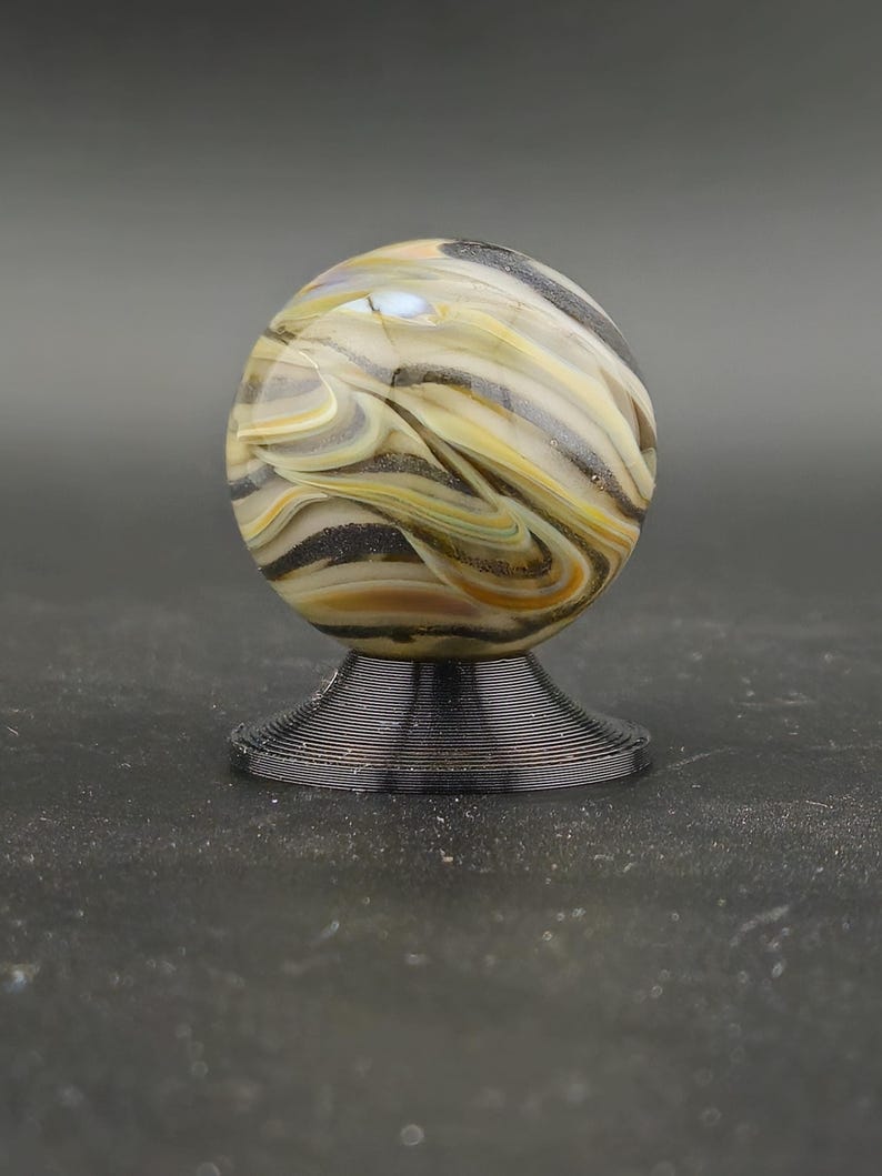 May include: A decorative glass marble with swirling patterns of tan, black, and yellow, resting on a black, wire-like stand. The marble is spherical and appears to be made of fused glass. The stand is circular and has a tiered design.