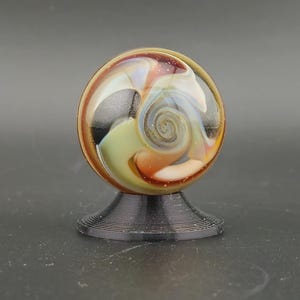 May include: A spherical glass marble featuring swirling patterns of amber, cream, and black with iridescent highlights, resting on a small, circular, black, ribbed plastic display stand.
