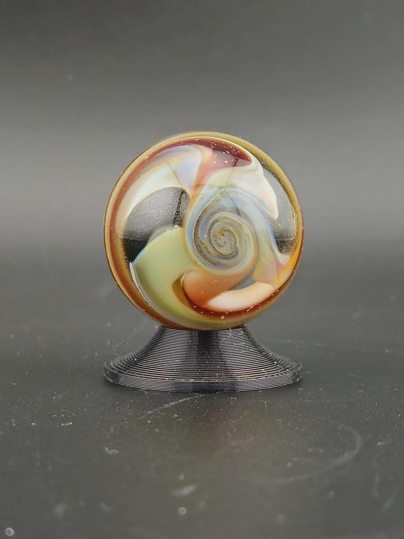 May include: A spherical glass marble featuring swirling patterns of amber, cream, and black with iridescent highlights, resting on a small, circular, black, ribbed plastic display stand.