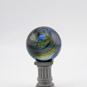 May include: A decorative glass marble with swirling blue, green, and gold colors, resting on a small, gray, classical-style pedestal. The marble has a spherical shape and a glossy finish, creating a visually appealing display.