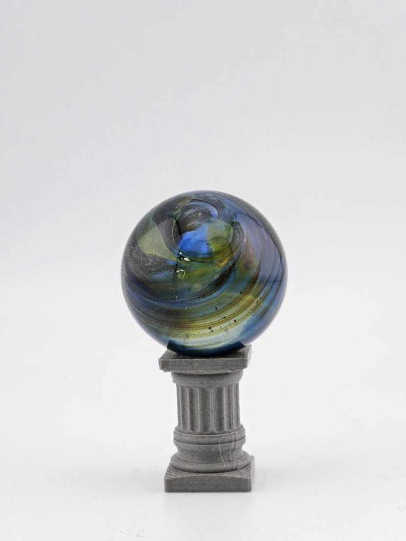 May include: A decorative glass marble with swirling blue, green, and gold colors, resting on a small, gray, classical-style pedestal. The marble has a spherical shape and a glossy finish, creating a visually appealing display.