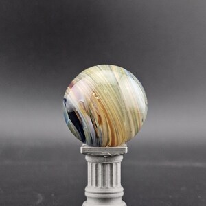 May include: A decorative glass sphere with swirling colors of gold, brown, and blue, resting on a small, gray, fluted column. The sphere has a polished, reflective surface. The column is a miniature replica of a classical architectural element.