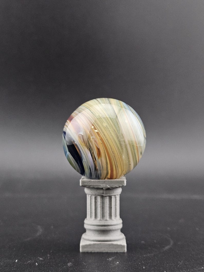 May include: A decorative glass sphere with swirling colors of gold, brown, and blue, resting on a small, gray, fluted column. The sphere has a polished, reflective surface. The column is a miniature replica of a classical architectural element.