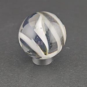 May include: A spherical glass marble featuring a clear base with swirling opaque white ribbons and dark, shimmering blue-black glitter inclusions throughout the interior. The marble is resting on a small, circular, dark gray support stand.