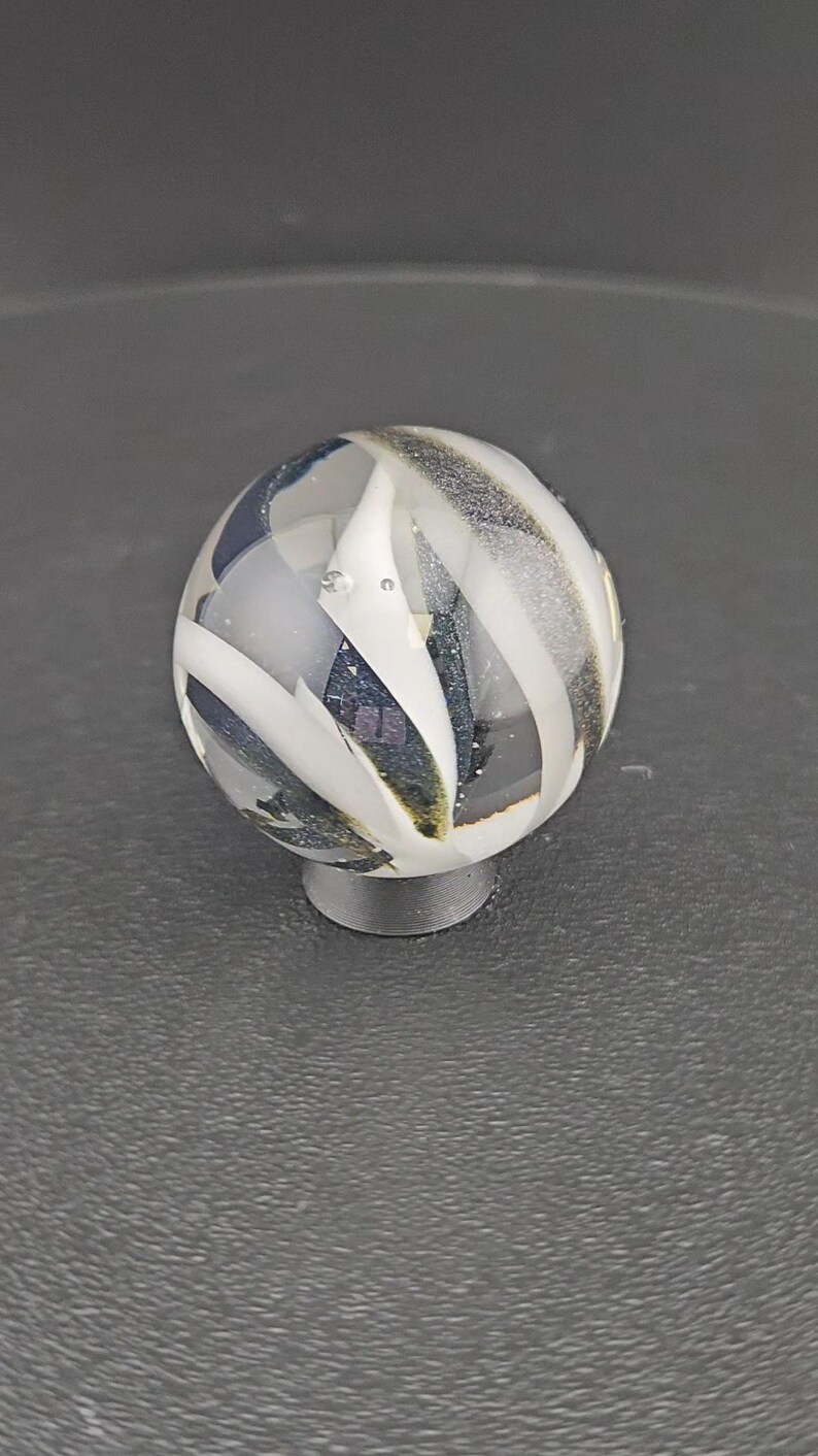 May include: A spherical glass marble featuring a clear base with swirling opaque white ribbons and dark, shimmering blue-black glitter inclusions throughout the interior. The marble is resting on a small, circular, dark gray support stand.