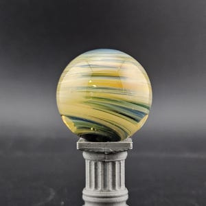 May include: A decorative glass marble with yellow, blue, and green swirls, resting on a small, gray, fluted column. The marble has a translucent quality, and the column has a classic architectural design.