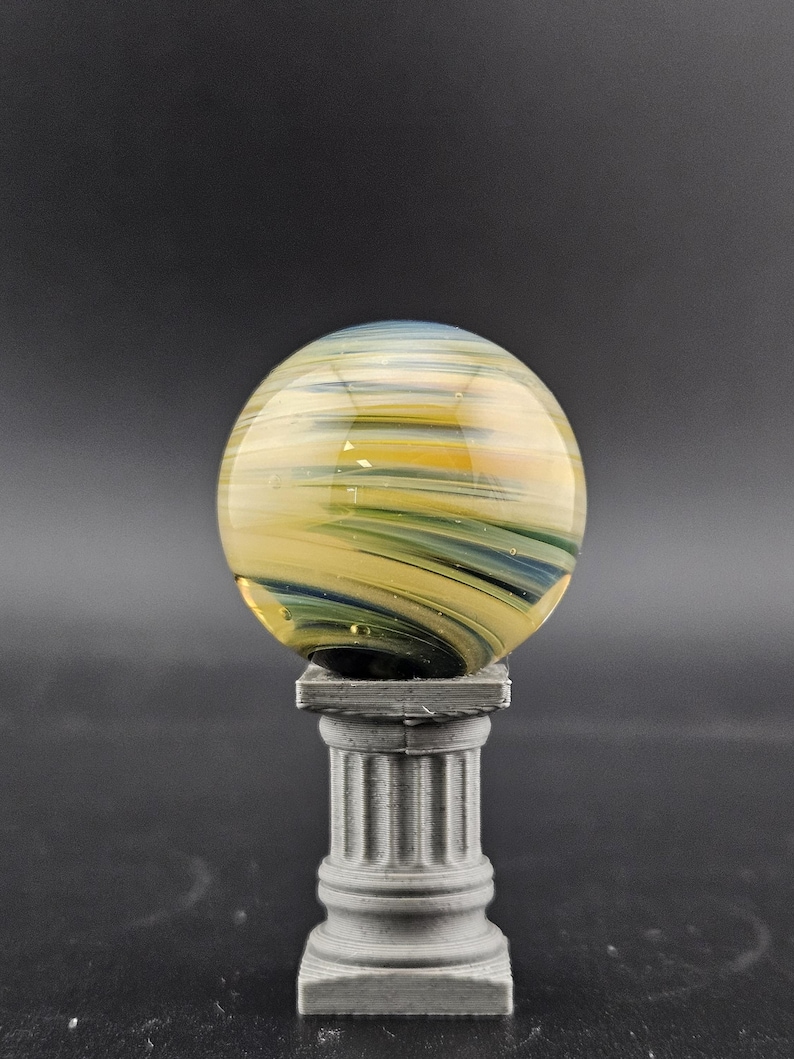 May include: A decorative glass marble with yellow, blue, and green swirls, resting on a small, gray, fluted column. The marble has a translucent quality, and the column has a classic architectural design.
