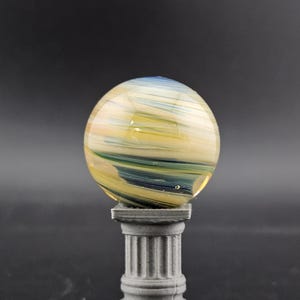 May include: A glass sphere with yellow, blue, and white swirls, resting on a gray, fluted column. The sphere has a translucent quality, and the column has a classical design. The background is a solid dark gray.