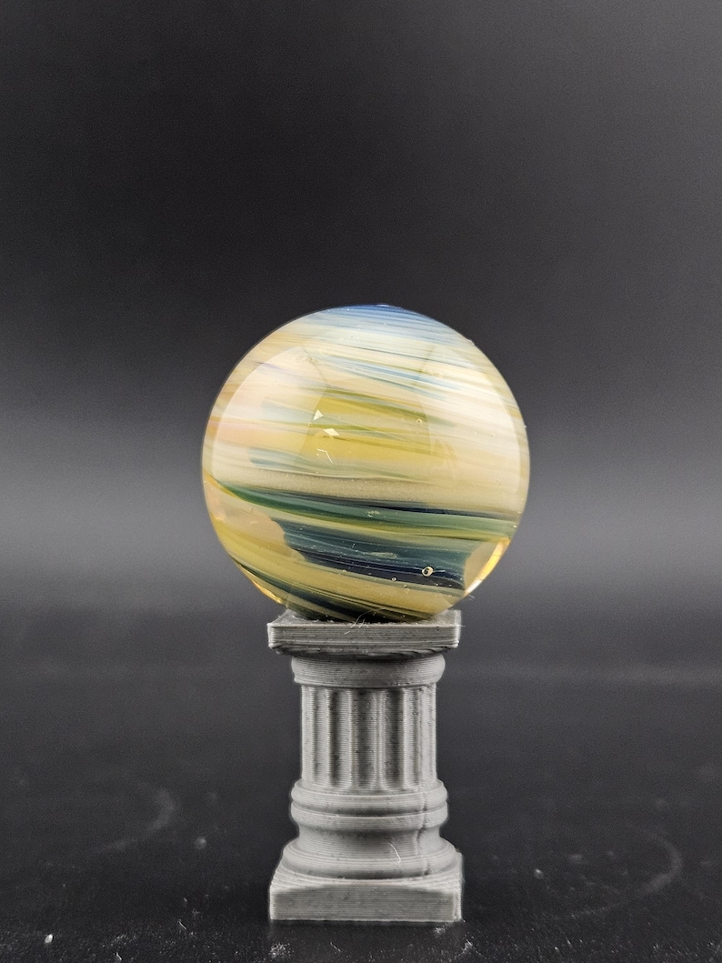 May include: A glass sphere with yellow, blue, and white swirls, resting on a gray, fluted column. The sphere has a translucent quality, and the column has a classical design. The background is a solid dark gray.