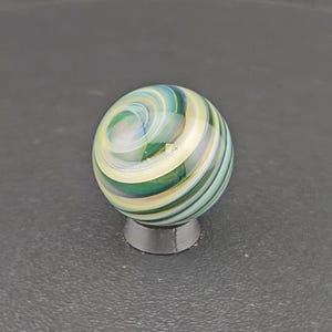 May include: A spherical glass marble featuring a swirling pattern of translucent green, cream, and light blue colors. The surface is smooth and polished, reflecting light across the intricate, layered bands that spiral around the center of the orb.