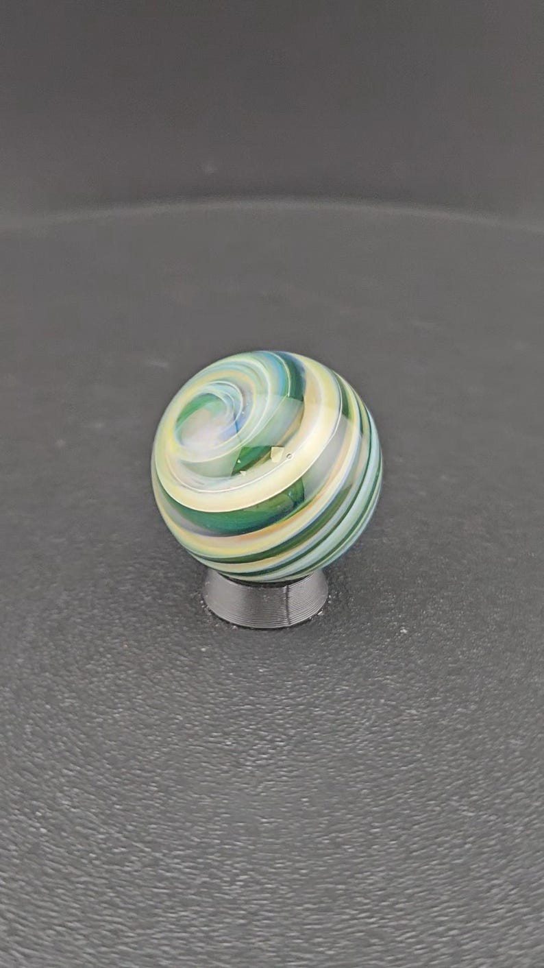 May include: A spherical glass marble featuring a swirling pattern of translucent green, cream, and light blue colors. The surface is smooth and polished, reflecting light across the intricate, layered bands that spiral around the center of the orb.