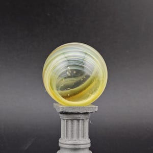 May include: A clear glass marble with yellow and green swirls, resting on a gray, fluted column pedestal. The marble has a spherical shape and is the focal point of the image. The pedestal has a square base.