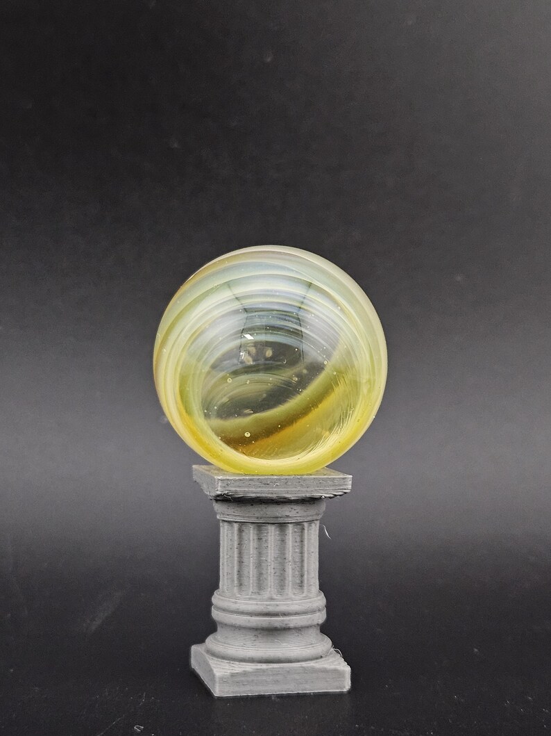 May include: A clear glass marble with yellow and green swirls, resting on a gray, fluted column pedestal. The marble has a spherical shape and is the focal point of the image. The pedestal has a square base.
