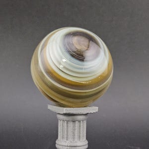 May include: A decorative glass marble with swirling patterns of brown, white, and gold, resting on a small, gray, classical-style pedestal. The marble has a glossy finish and is the focal point of the image.