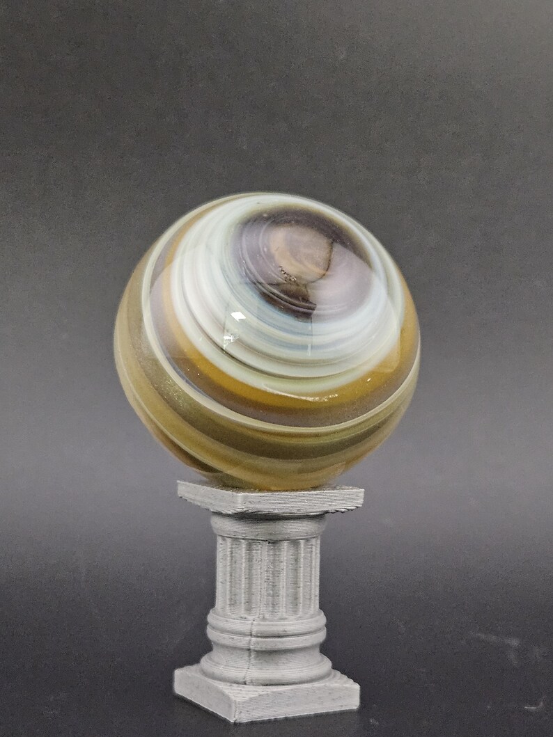 May include: A decorative glass marble with swirling patterns of brown, white, and gold, resting on a small, gray, classical-style pedestal. The marble has a glossy finish and is the focal point of the image.