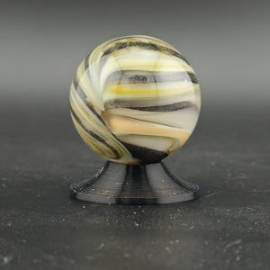 May include: A decorative glass marble with a swirl pattern of yellow, black, and white. The marble sits on a black, ridged base. The background is a solid grey.