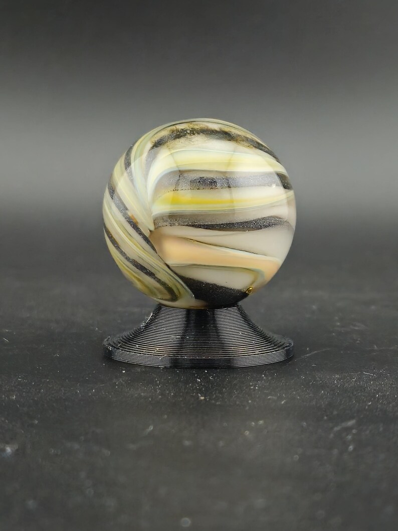 May include: A decorative glass marble with a swirl pattern of yellow, black, and white. The marble sits on a black, ridged base. The background is a solid grey.