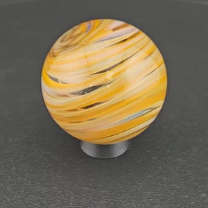 May include: A spherical glass marble featuring a swirling pattern of translucent yellow, orange, and white bands. The orb has a smooth, polished surface with a glossy finish, displaying intricate, layered color streaks that wrap around the sphere.