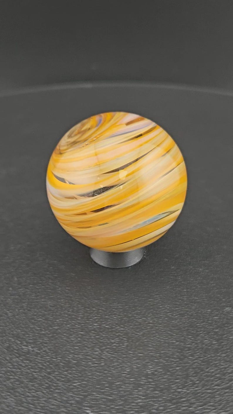 May include: A spherical glass marble featuring a swirling pattern of translucent yellow, orange, and white bands. The orb has a smooth, polished surface with a glossy finish, displaying intricate, layered color streaks that wrap around the sphere.