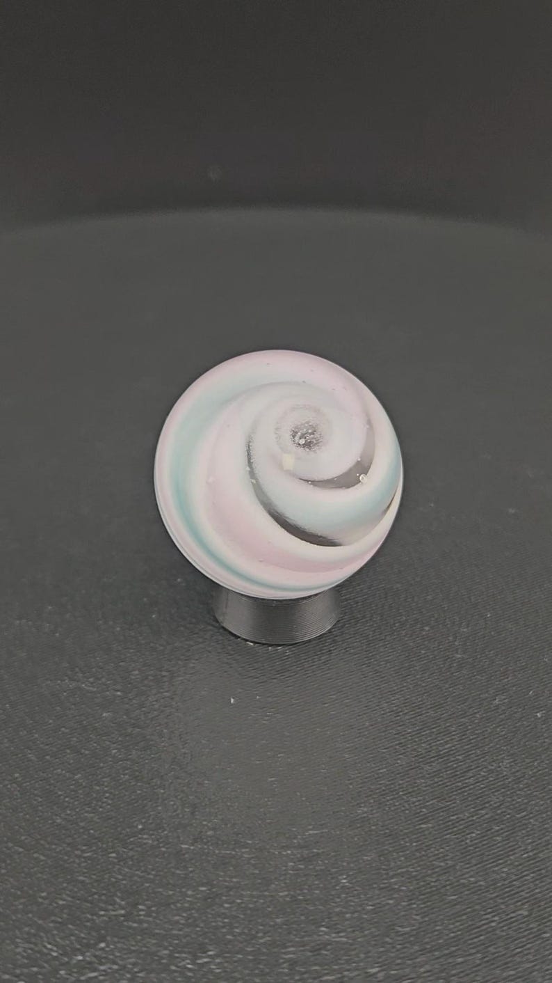 K&ouml;nnte beinhalten: Eine kugelf&ouml;rmige Glasmurmel mit einem wirbelnden Muster in Pastellrosa, Hellblau und Wei&szlig;. Die Oberfl&auml;che der Murmel ist glatt und gl&auml;nzend, wobei das Spiralmuster zu einem zentralen Punkt an der Oberseite der Kugel hin zusammenl&auml;uft.
