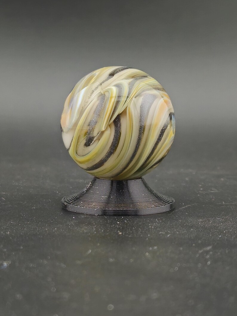 May include: A decorative glass marble with swirling patterns of yellow, black, and white, on a black, spiral-shaped stand. The marble's design resembles natural stone. The stand is approximately 2.5 cm in diameter.