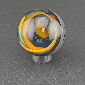 May include: A spherical glass marble featuring swirling internal patterns of vibrant orange, yellow, green, and blue colors set against a clear glass base. The marble is resting on a small, circular, dark gray stand, showcasing its smooth surface.