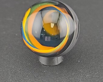 May include: A spherical glass marble featuring swirling internal patterns of vibrant orange, yellow, green, and blue colors set against a clear glass base. The marble is resting on a small, circular, dark gray stand, showcasing its smooth surface.