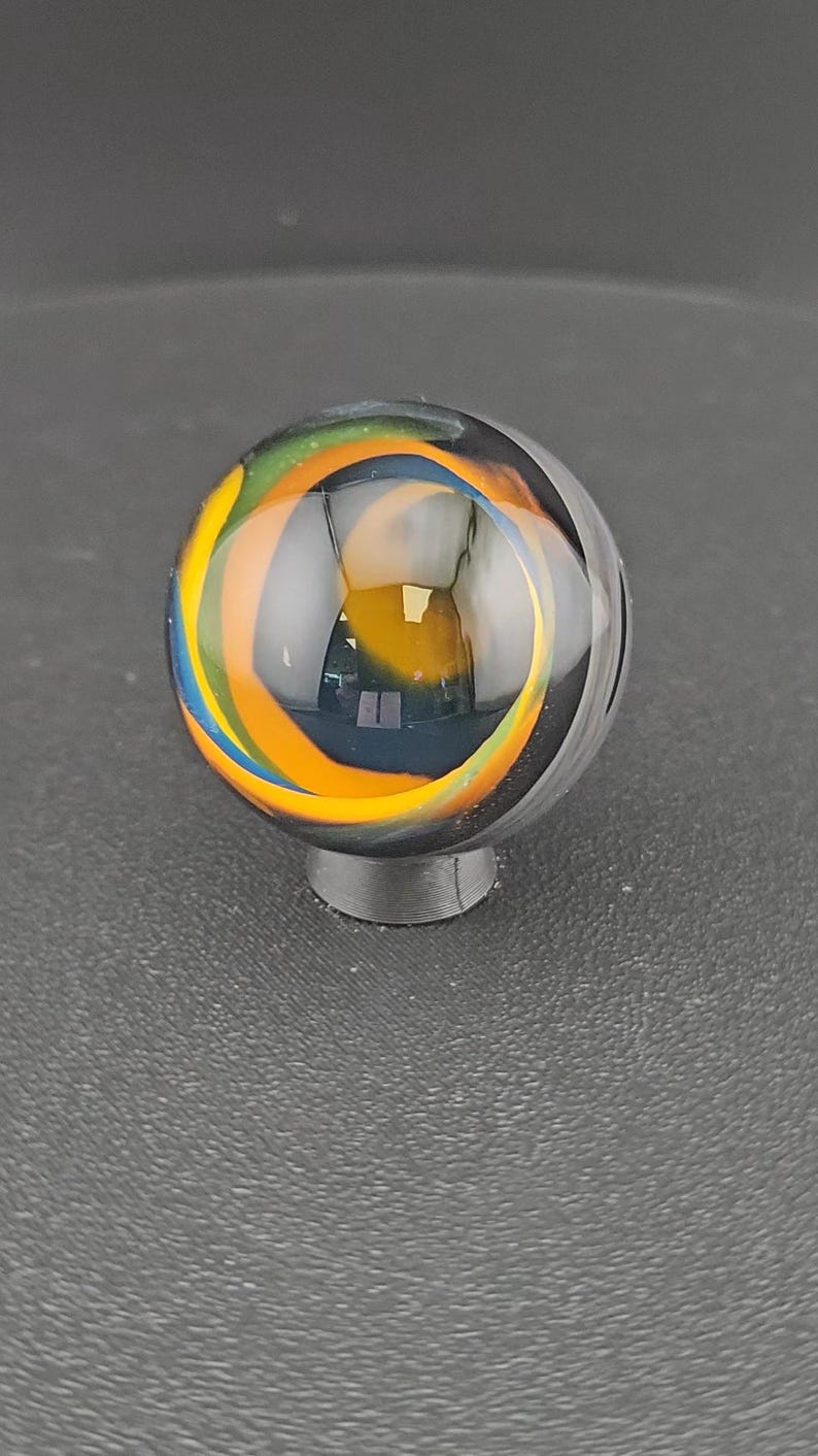 Op de afbeelding: Een bolvormige glazen knikker met wervelende interne patronen in levendige oranje, gele, groene en blauwe kleuren op een basis van helder glas. De knikker rust op een kleine, ronde, donkergrijze standaard en toont het gladde oppervlak.