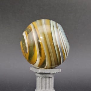 May include: A large, colorful glass marble with yellow, white, and brown swirling patterns. The marble is displayed on a small, gray, classical-style pedestal. The background is a solid, dark gray.
