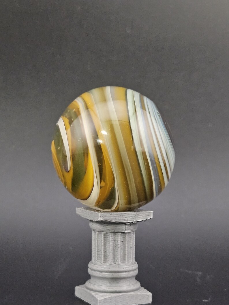 May include: A large, colorful glass marble with yellow, white, and brown swirling patterns. The marble is displayed on a small, gray, classical-style pedestal. The background is a solid, dark gray.