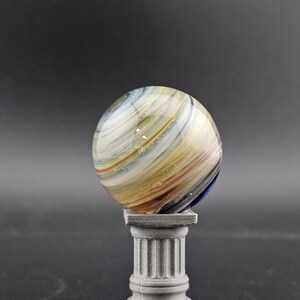 May include: A colorful glass marble with swirling patterns of white, yellow, and blue rests on a small, gray, classical-style pedestal. The marble's surface is reflective, and the pedestal has a fluted column design.